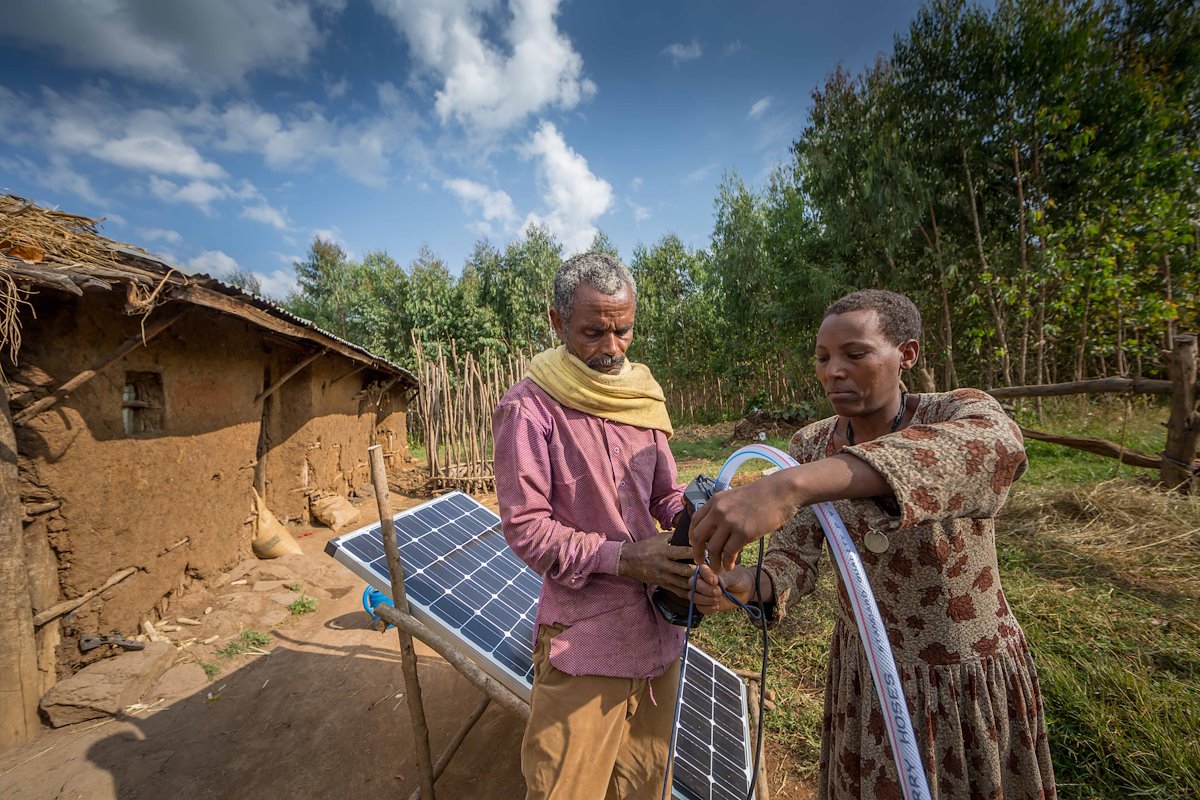 A man and woman setting up a solar panel next to a mud hut
