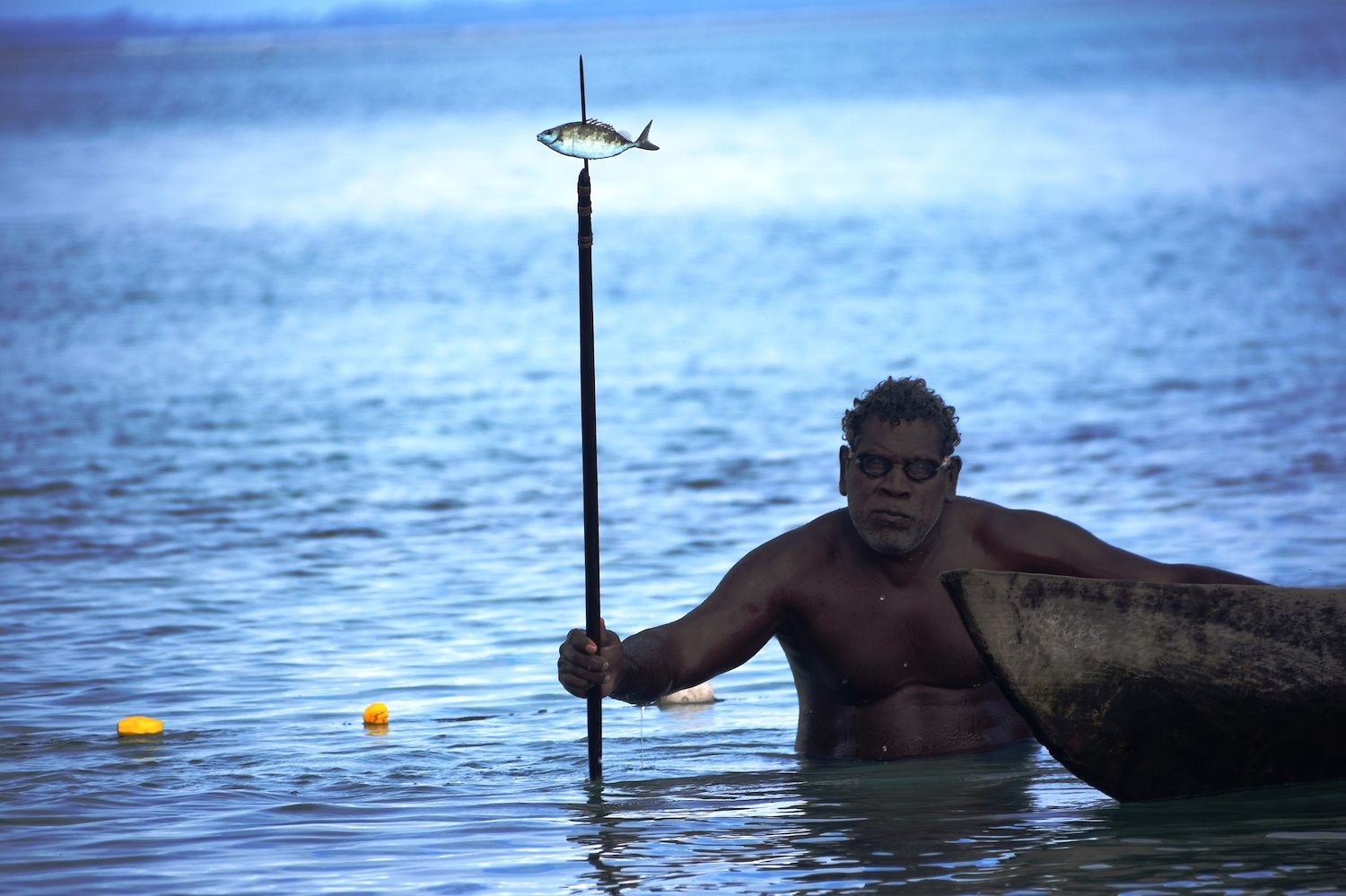 Man with goggles on holding long spear with small fish wading through ocean