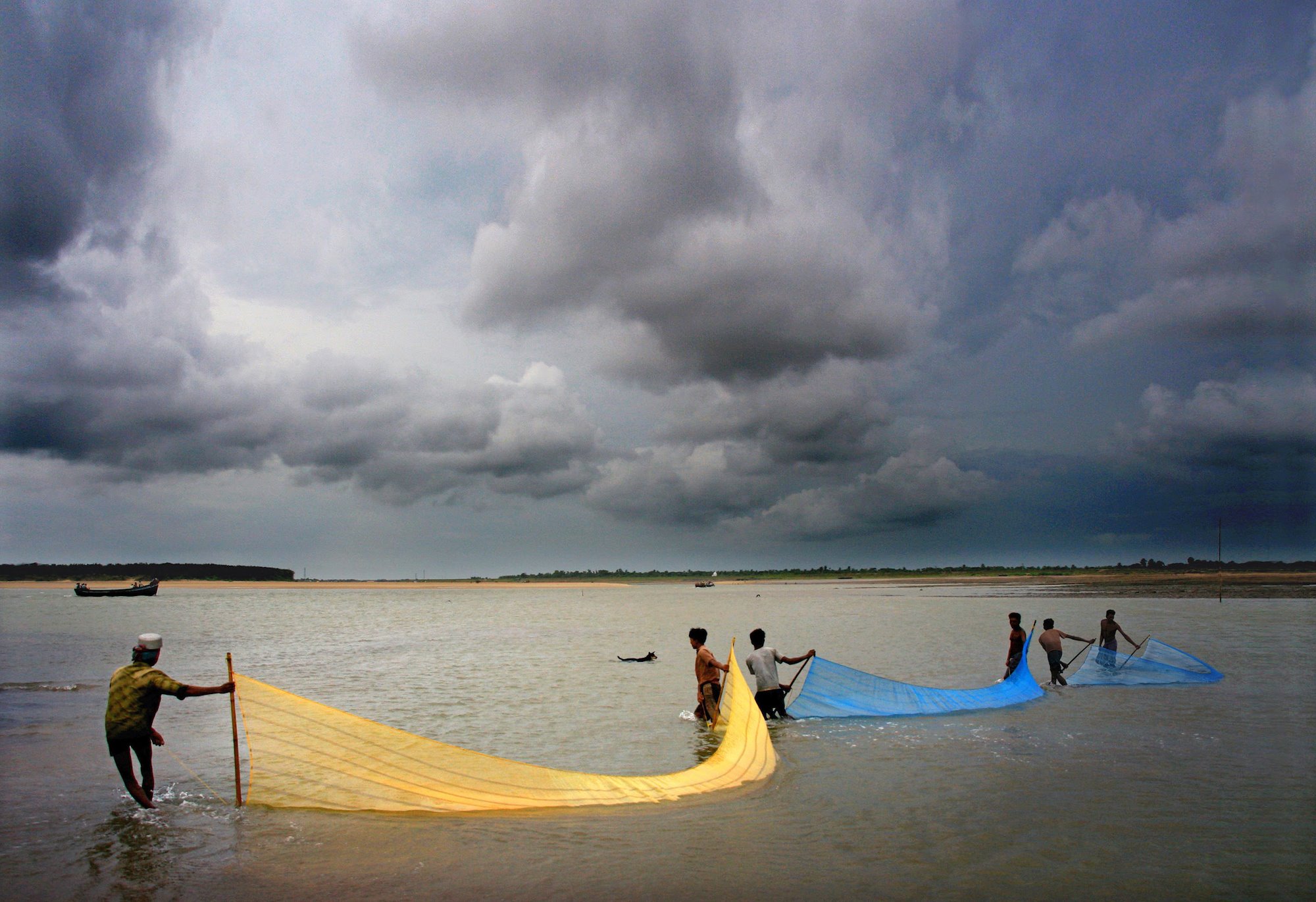 People pulling fishing nets into the ocean in front of storm clouds