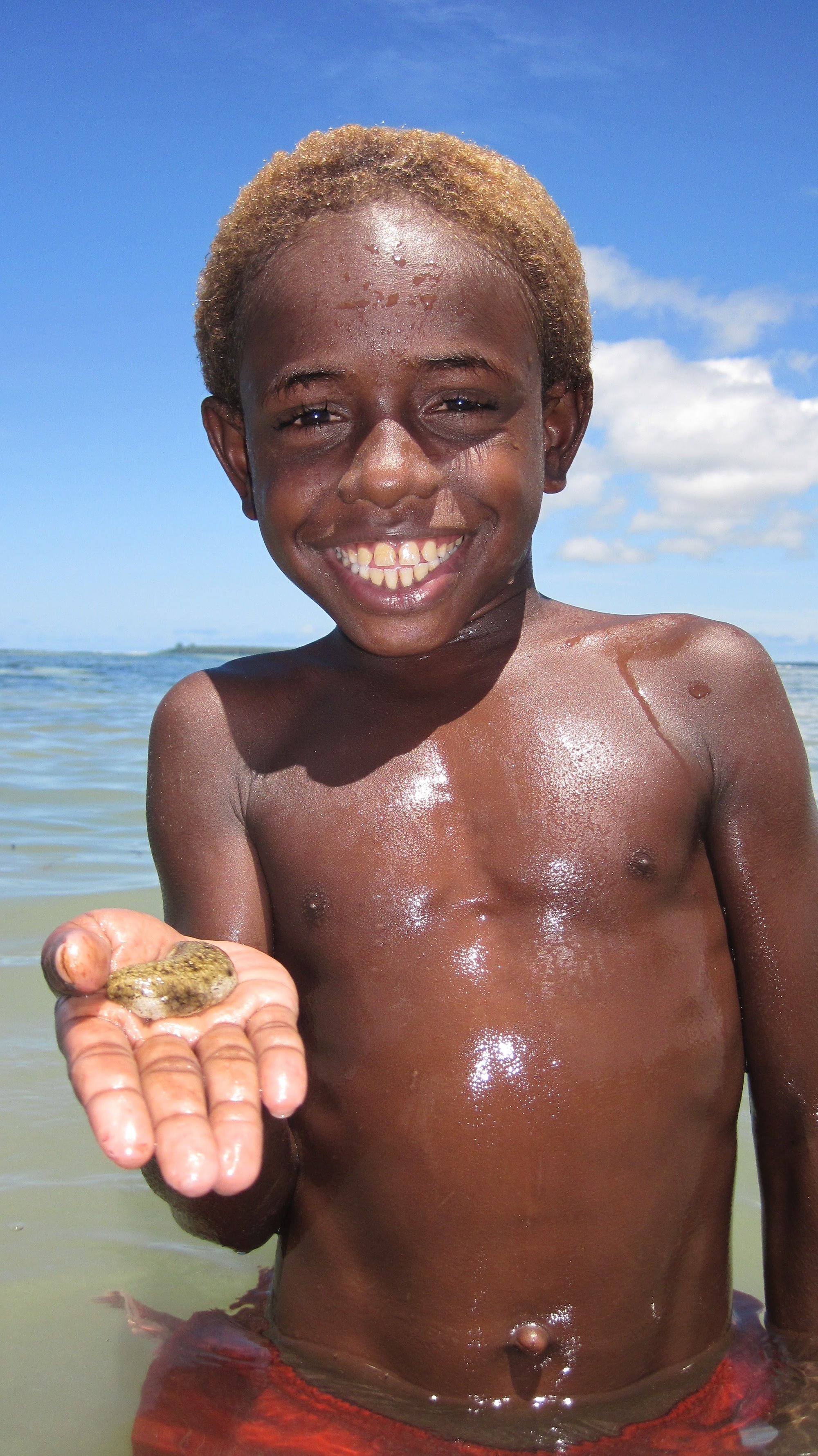 Young boy from PNG holding a small sea cucumber in the palm of his hand, standing in the ocean