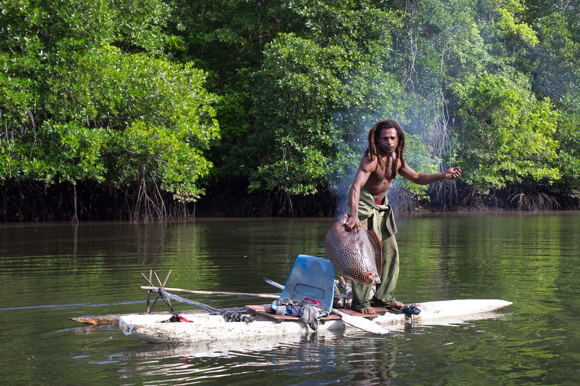 Man with long dreadlocks standing on paddle board holding large eel/fish