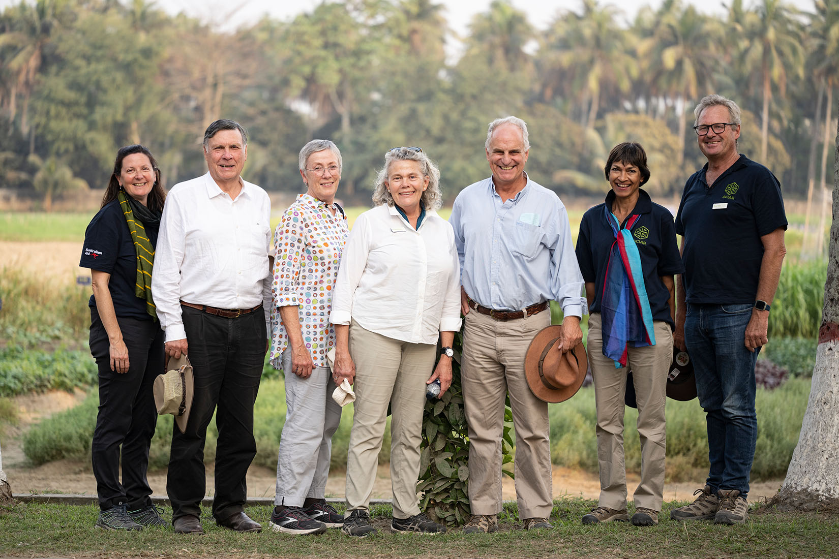 Seven people standing side by side, smiling at the camera, with trees and vegetation blurred in the background.