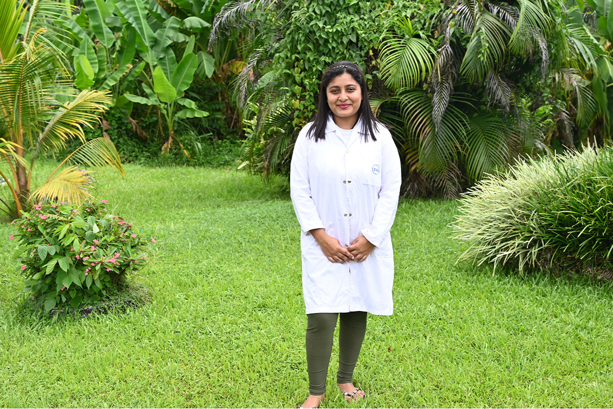 A woman in a white lab coat standing on a green lawn, with vegetation in the background.