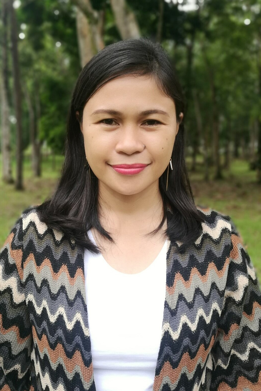 A woman with dark hair in a striped cardigan, with trees blurred in the background.