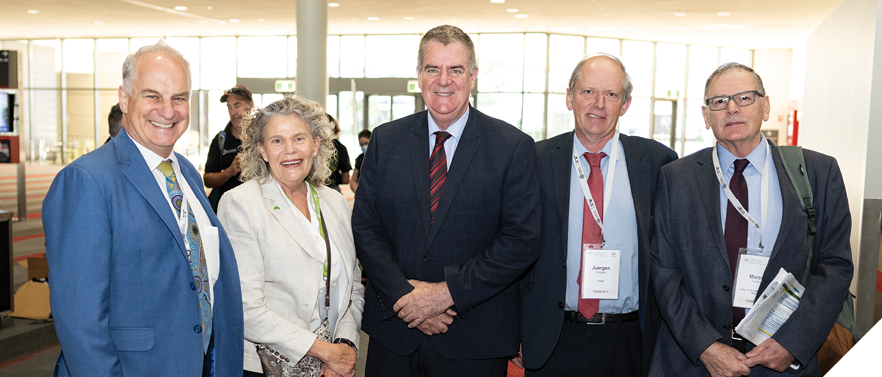 Four men and suits and ties and a woman wearing a white dress jacket and black pants pose for the camera. Some of them are wearing name tags. 