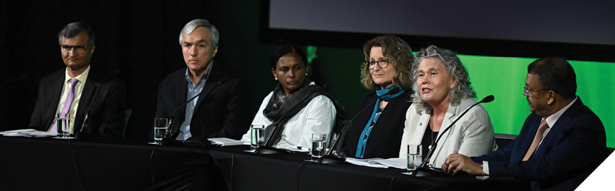 A group of three men and three women sit at a table on a stage with microphones in front of them. A woman in the middle, wearing a white dress jacket, is speaking. 