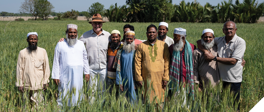 A group of 11 men standing in a row in a waist-high field. Some of them have their arms around each other’s shoulders. Several men are wearing hats and about half of the men have beards. 