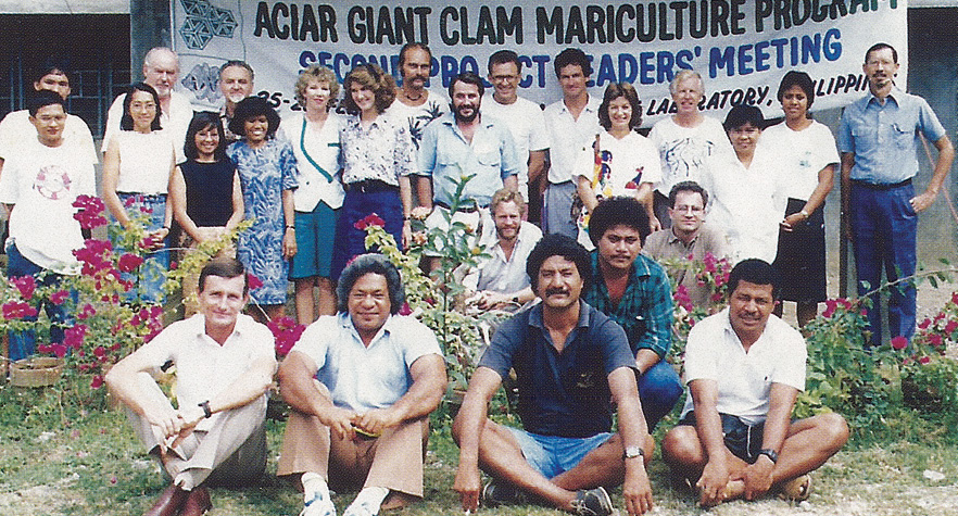 A group of 24 men and women sitting and standing in front of a sign that says ACIAR Giant Clam Mariculture Project. 