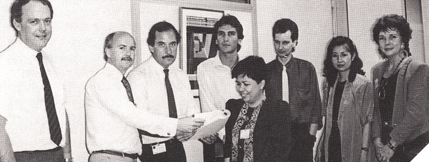 Black-and-white image of eight men and women. They are dressed formally, the men in ties and white shirts and the women in dress jacket. One of them in is holding out of book towards one of the women. 