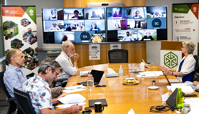 A group of men and women sitting around a board table. In front of them is a large screen showing a video conference call, with the remote attendee’s faces on the screen. 