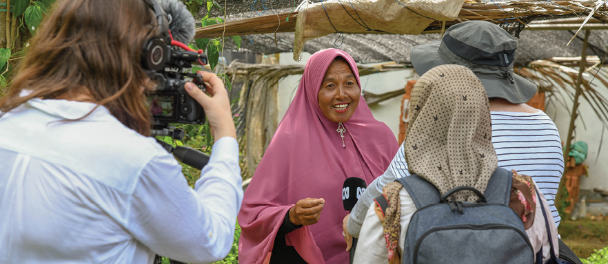 A woman in a pink headscarf is smiling and talking into a microphone being held buy two people in front of her, whose backs are towards the camera. There is also a video camera person to the left, and their back is also towards the camera. 