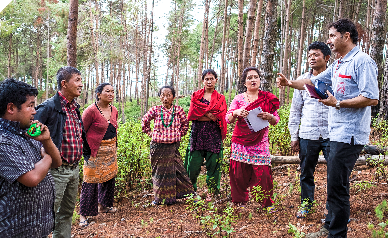 A group of people standing in a circle a forested area. They are listening to one man in the group speak. 