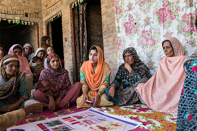 A group of women wearing colourful clothes are sitting on the floor looking at a poster on the ground. 