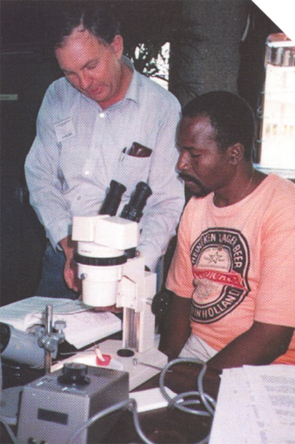 A man standing beside another man sitting at a large microscope. They are both looking at a book, which is open beside the microscope. 