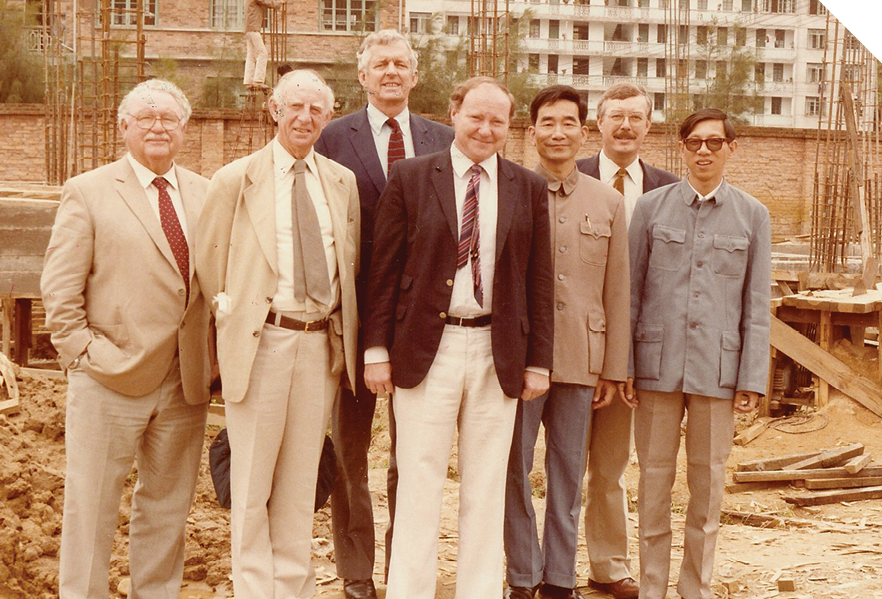 A group of seven men, most of whom are wearing suits and ties, are standing together on what looks like a construction site. The two men who are not wearing suits and ties are wearing formal button-up jackets. 