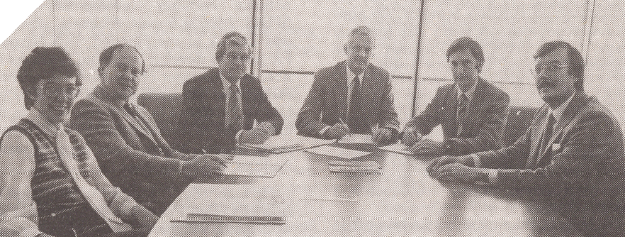 A black-and-white photo of five men in suits and one woman sitting around a board table. Their hands are on the table and they are holding pens, and there is paper scattered across the table.