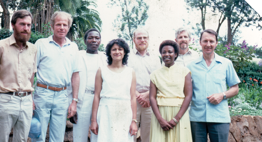 Six men and two women stand in a group together. They are all wearing pastel-coloured clothing. There are several tall tropical-looking trees in the background. 
