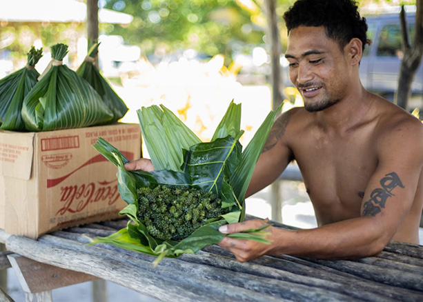A man wraps seaweed in large green leaves. He is sitting at a wooden table. Next to him is a cardboard box with wrapped seaweed in it. 