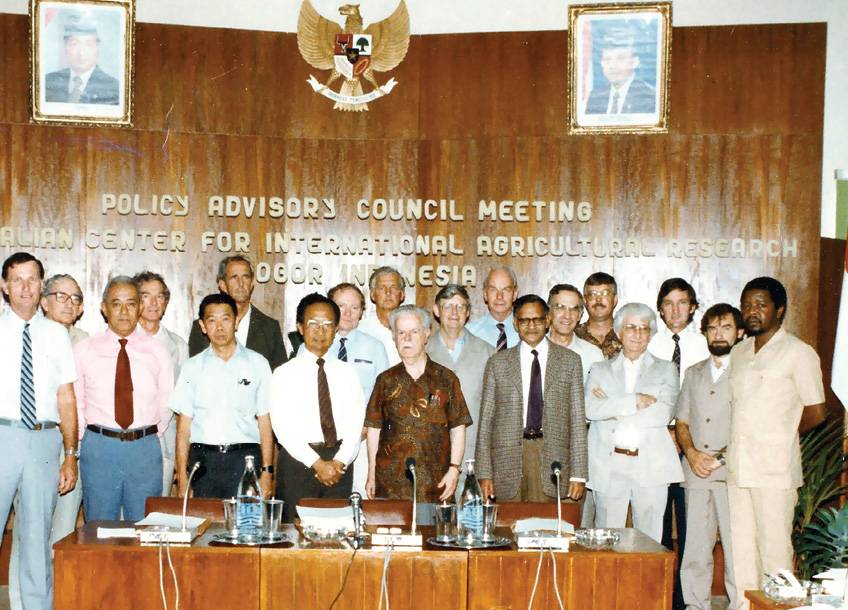 A group photo of 17 men. The men are dressed semiformally, most are wearing ties or jackets. They are standing behind a desk that has several microphones on it. Behind them is a wooden board with the text Policy Advisory Council Meeting written on it.