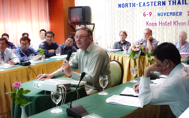 A group of people sitting at conference tables. The tables are arranged in a U-shape, and in the centre of the U-shape is a man sitting at a small table with a projector. He is talking into a microphone. 