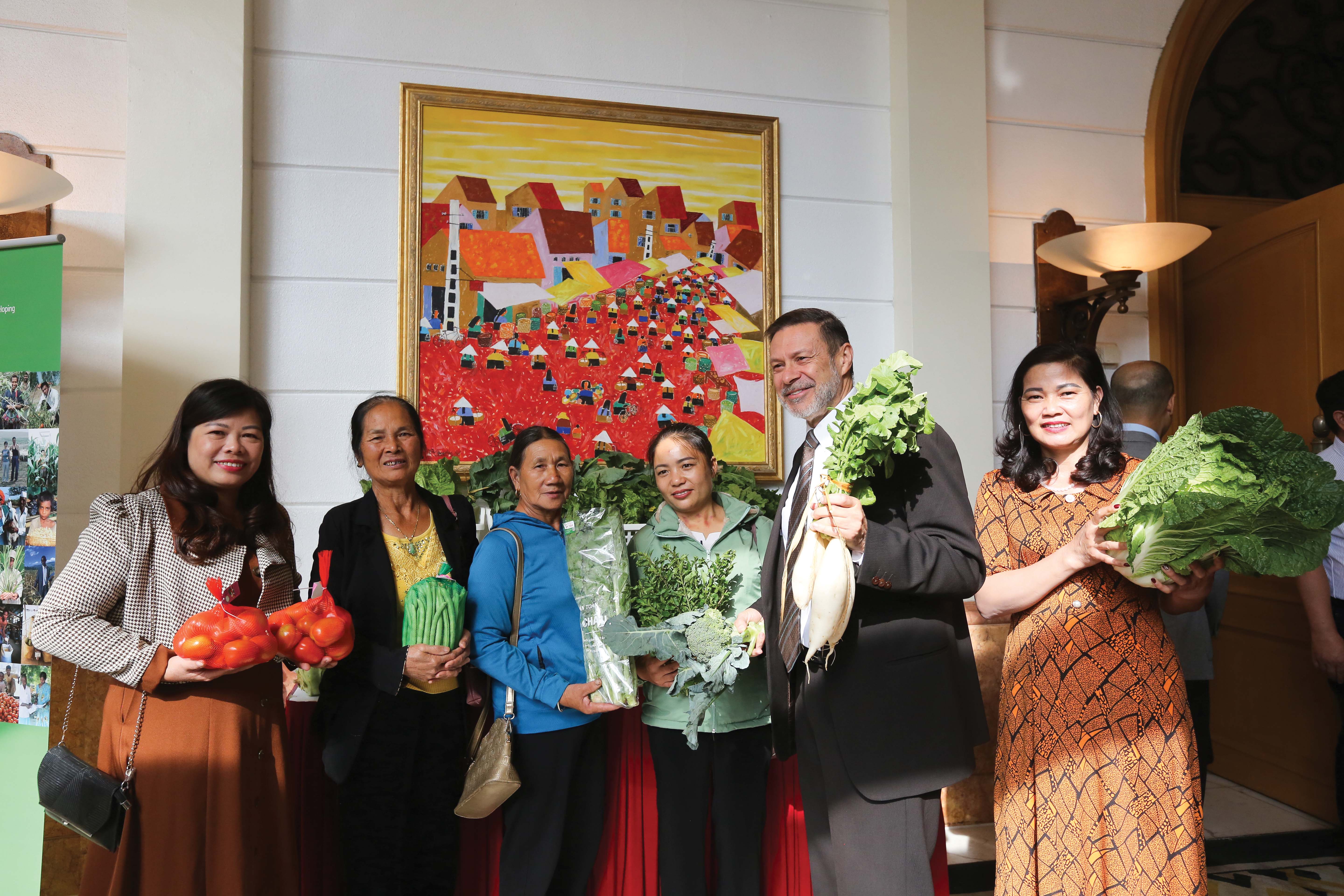 a group of women and a man standing in front of a colourful painting. They each hold a different vegetable, and pose for the photo.