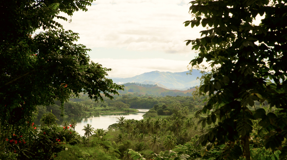 A wide shot of Kiribati shows a water body surrounded by lush green vegetation, with mountains in the distance.
