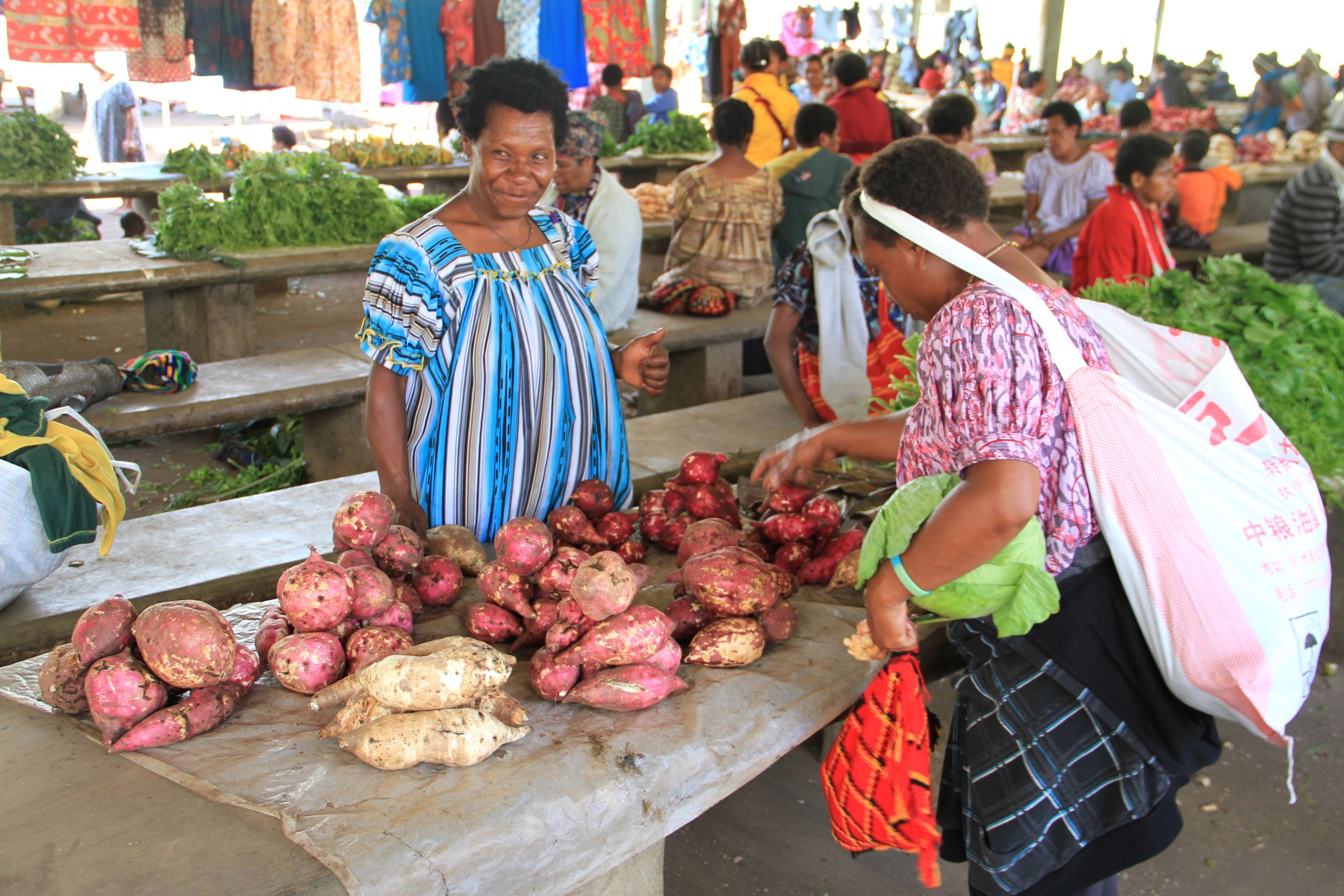 A customer browses the market stall of a woman offering sweetpotatoes for sale. 