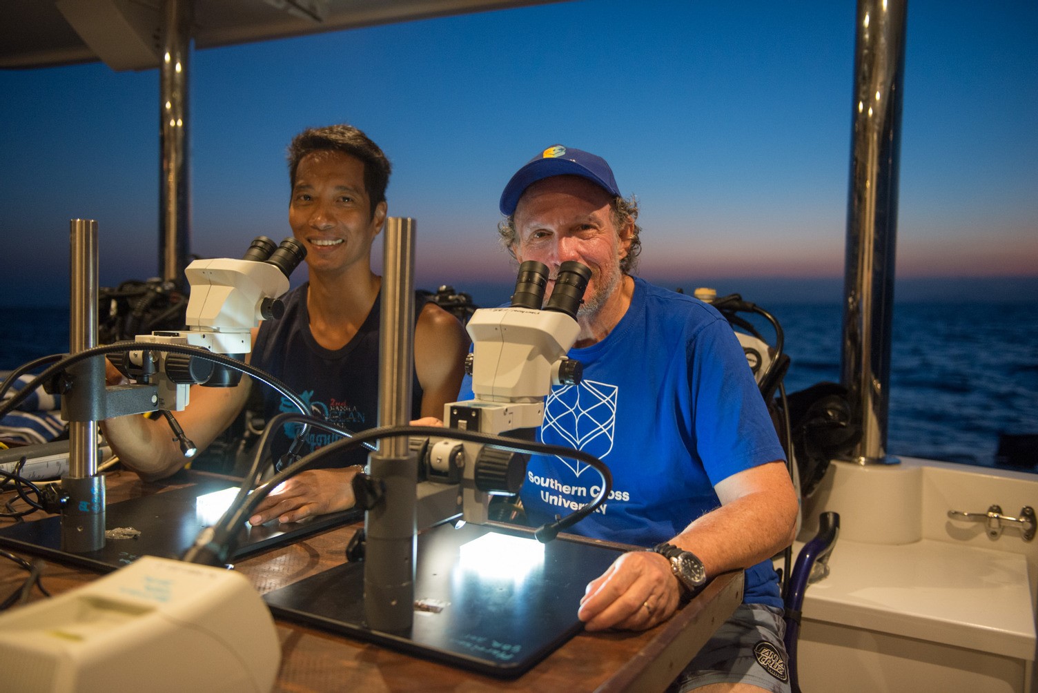 Two men sitting behind microscopes smiling with ocean in the background