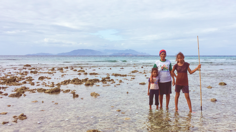 A woman and two young girls in ankle-deep, clear, sea water in Timor-Leste. The eldest girl holds a spear in her right hand..