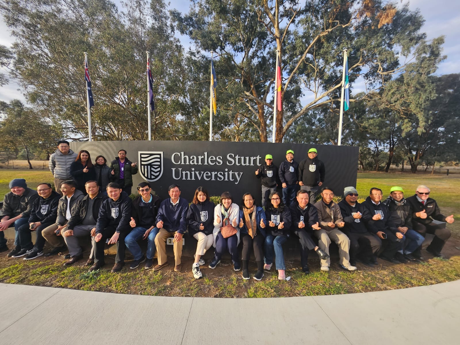 Large group of people sitting and standing outside in front of a large sign that say Charles Sturt University.