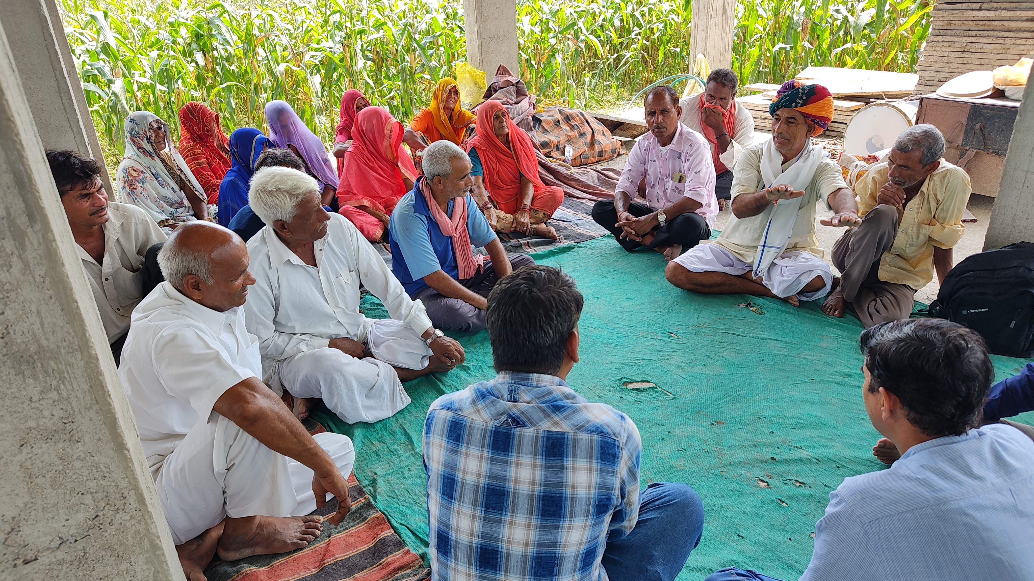 A group of adults sitting on the floor in a semi-circle under a shelter, engaging in a discussion. Some are wearing traditional Indian attire.