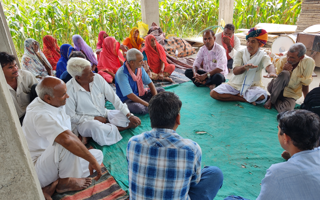 Group of people sitting in a circle on a blue mat. Ladies wearing colourful headscraves