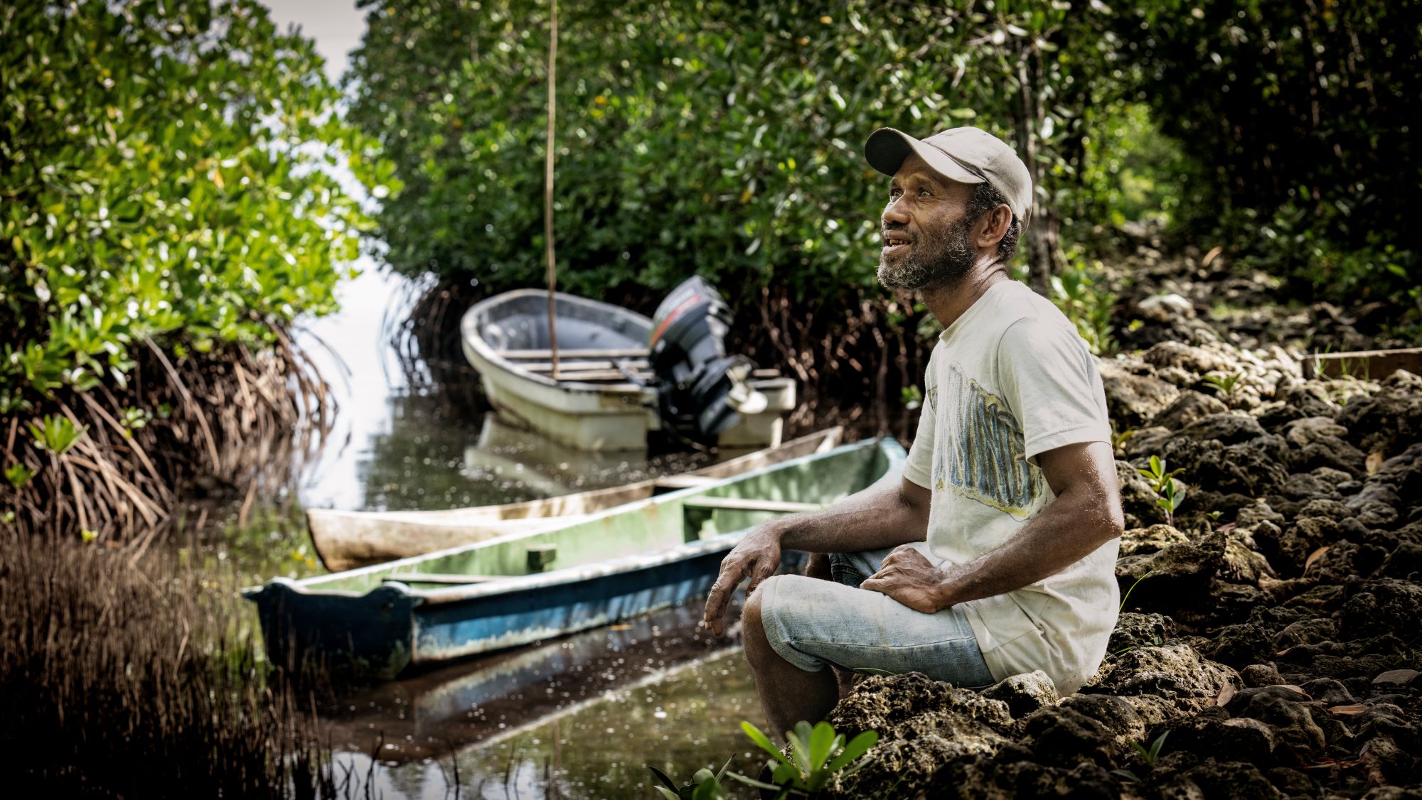 Fisherman sitting on rocky ground next to mangrove-lined water with two small canoes nearby. 