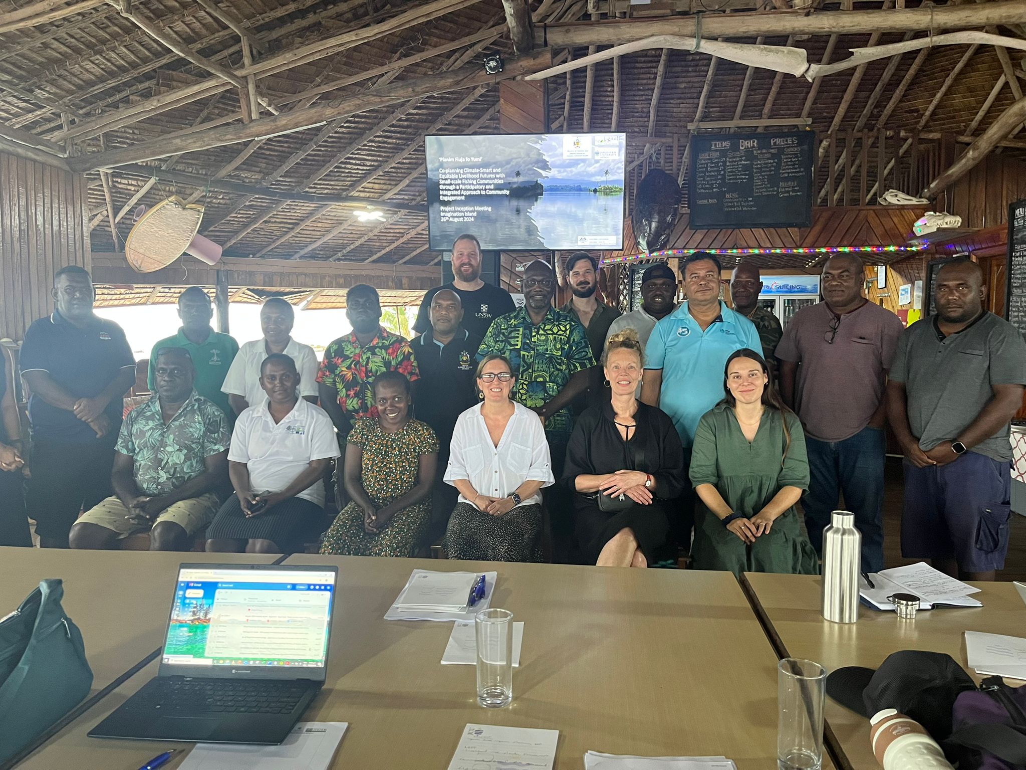 Two rows of people facing the camera sit in front of a screen.