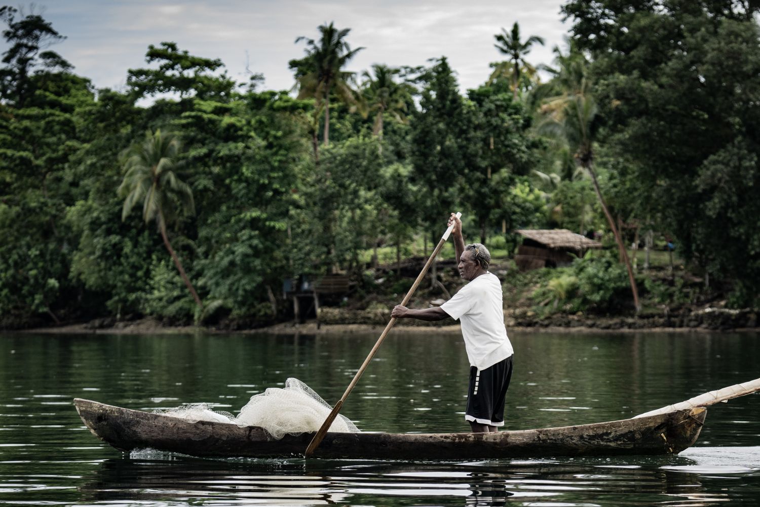A person stands in a wooden canoe, paddling across water with tropical trees in the background.