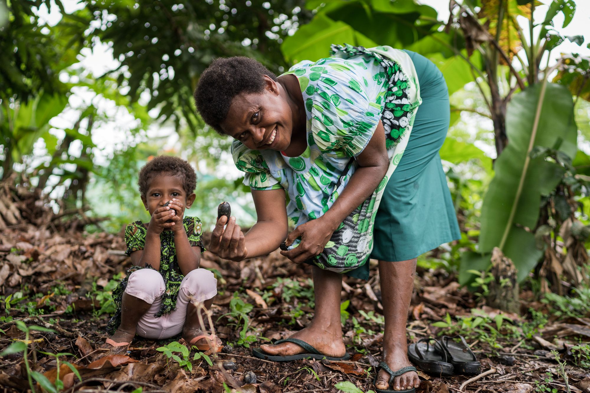 An adult and child in a tropical outdoor setting; the child sits while the adult smiles sand holds a dark nut in her hand