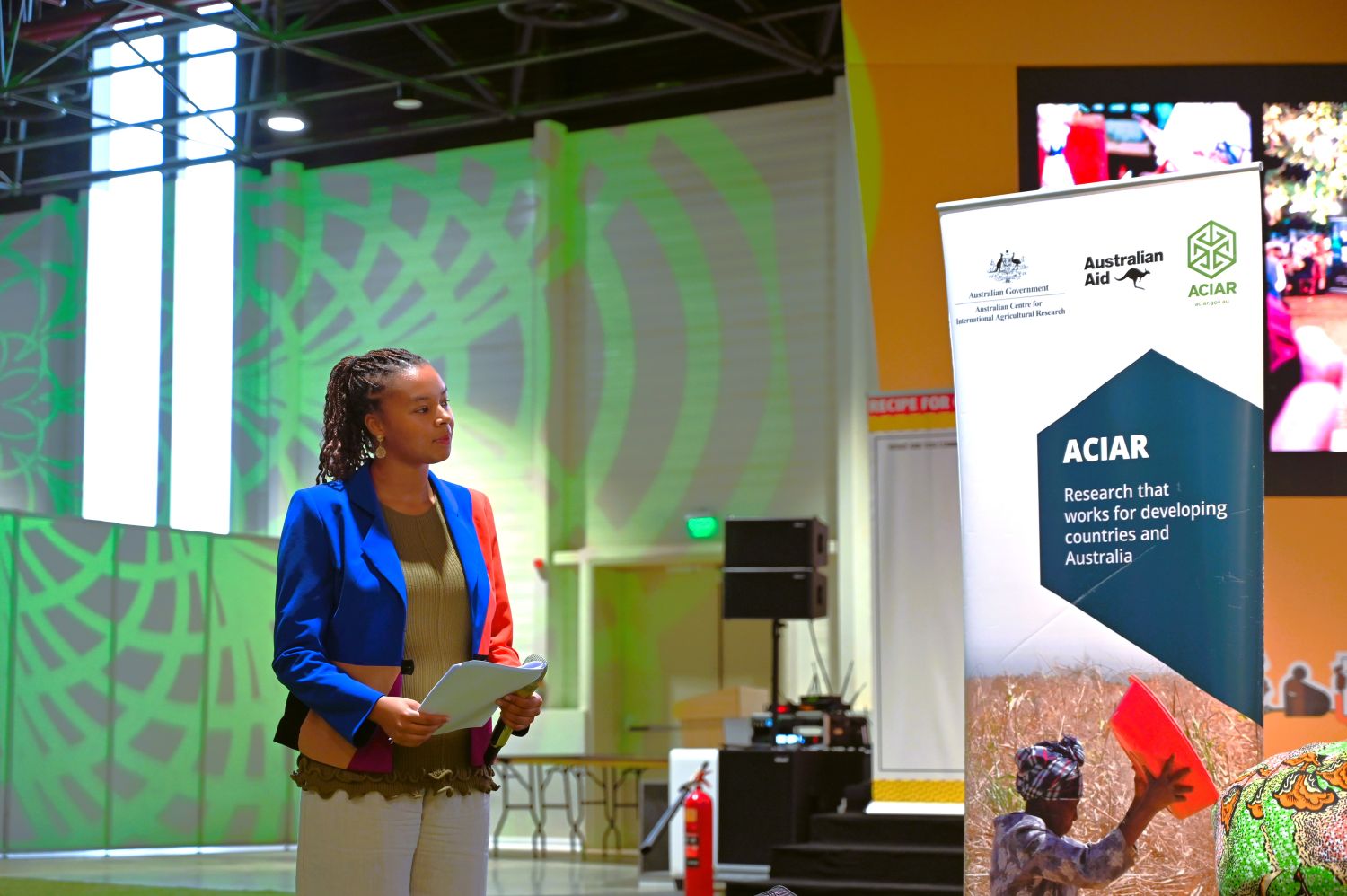 A women speaker holds notes on stage beside an ACIAR pull-up banner
