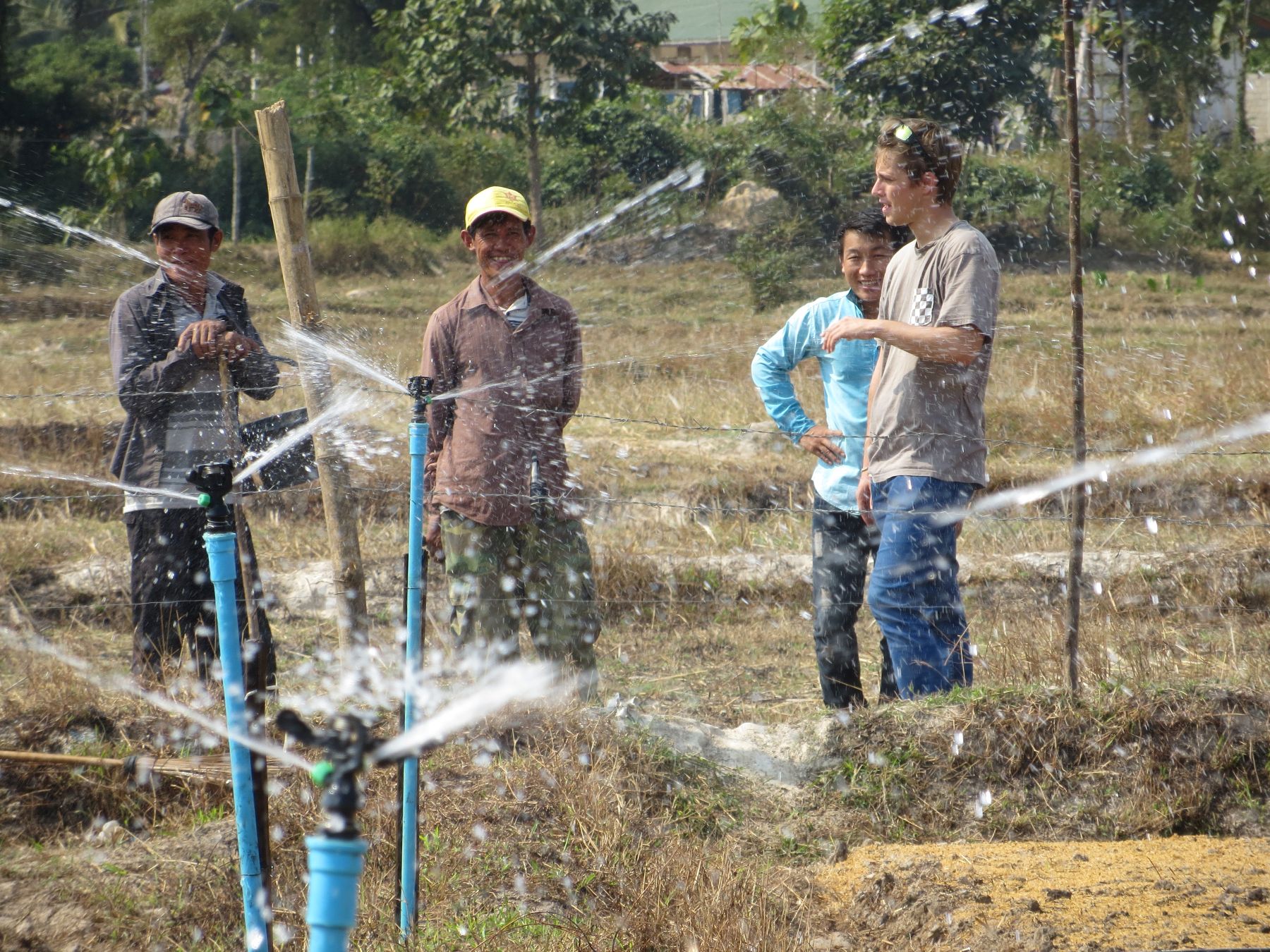 Four people stand in a field observing a working irrigation sprinkler. 