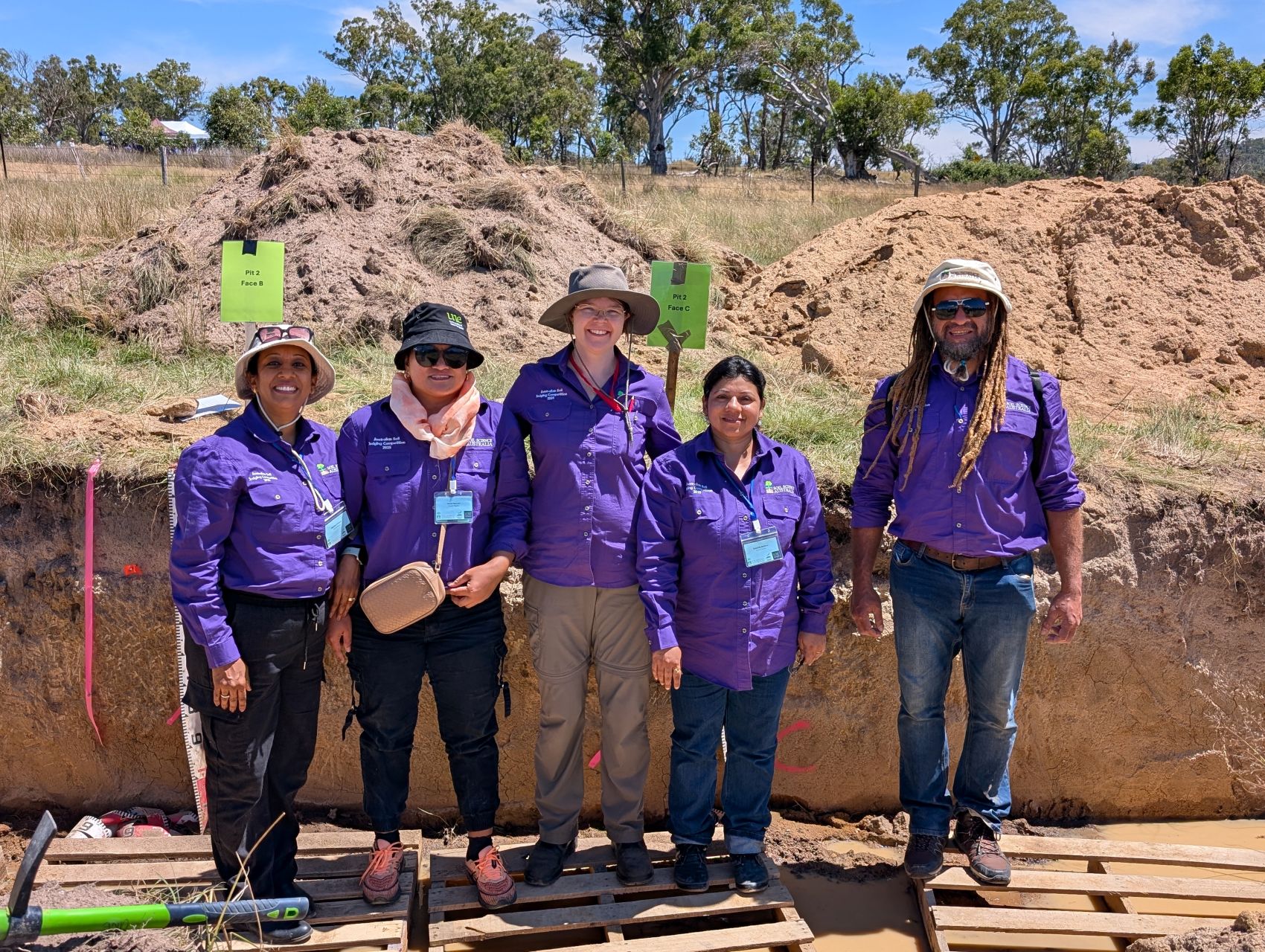 A group of people wearing purple shirts stand in a row in front of a carved our wall of soil. 