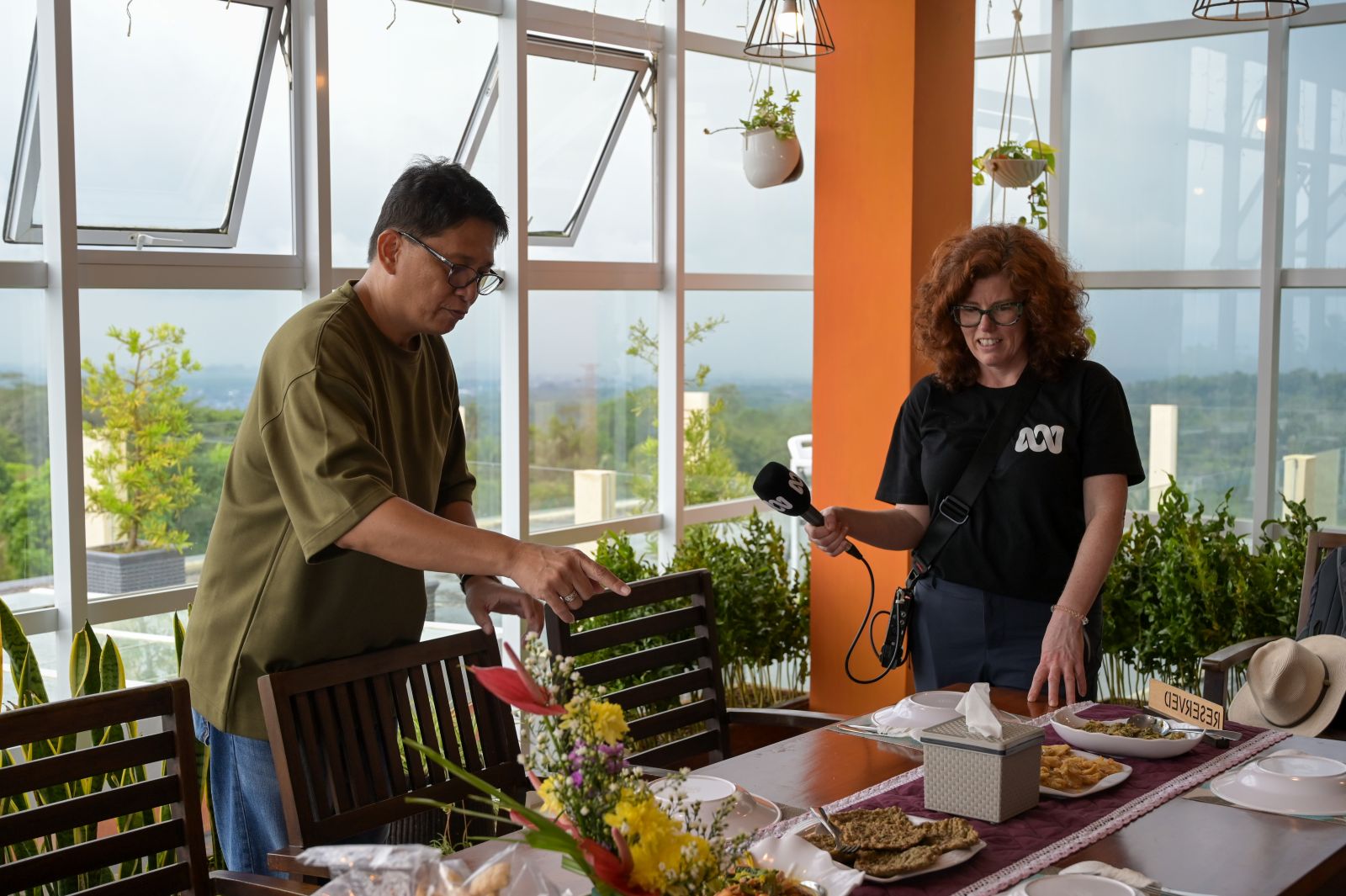 Two people stand near a dining table set with plates of food and a floral centerpiece in a bright room with large windows and hanging plants.