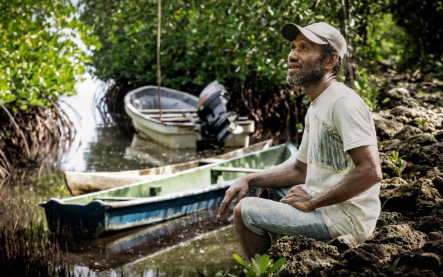 Person sitting on a rocky shore beside small boats in a mangrove-lined waterway.