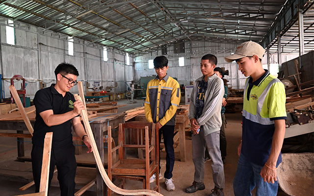 A group of people stand inside a woodworking workshop as one person demonstrates a curved wooden piece, with tools and timber visible in the background.