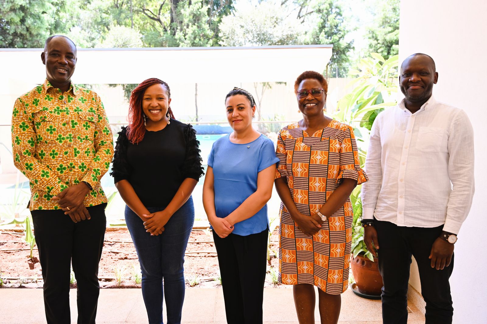 Five people stand side by side outside under cover, wearing a mix of patterned and solid-colour clothing and smiling at the camera.