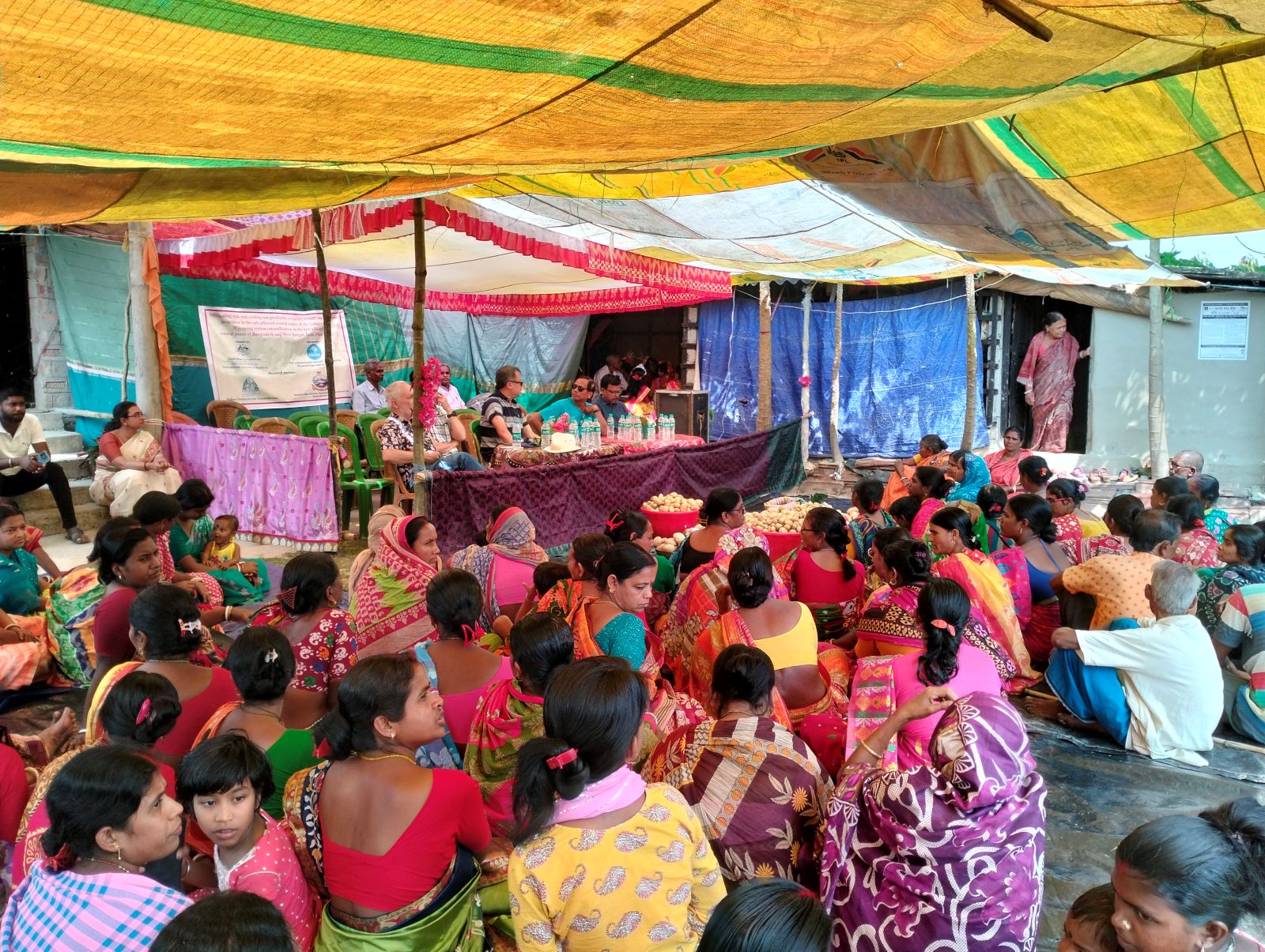An outdoor community gathering with participants wearing brightly coloured clothing beneath fabric shelters.