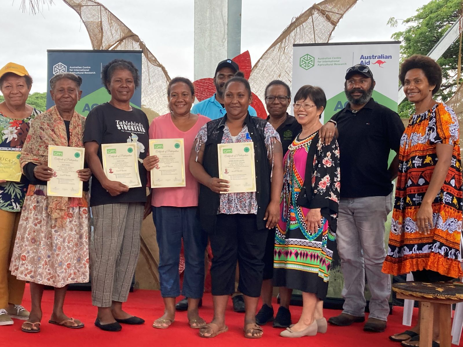Group of participants standing on a stage holding certificates, with ACIAR and Australian Aid banners behind them.