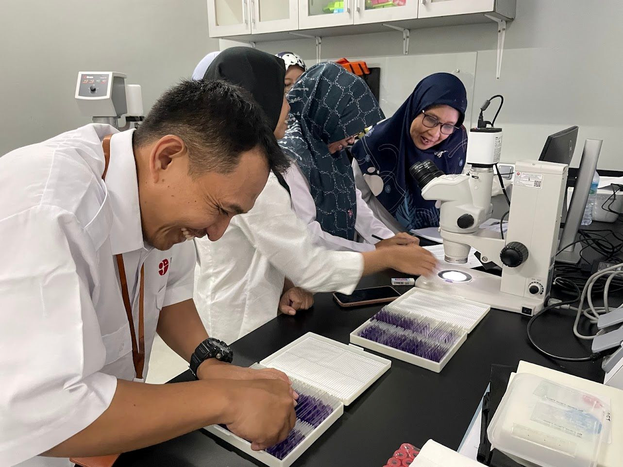 Researchers examine prepared samples at a laboratory bench using a microscope and sample trays.