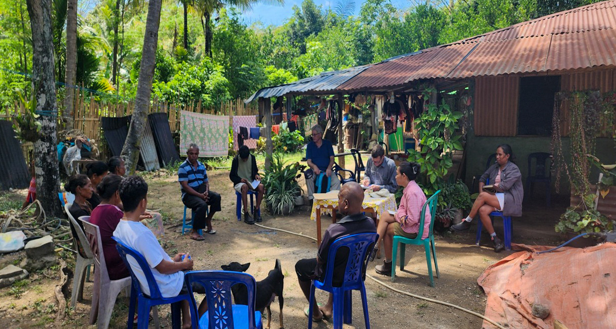 Community members sit in a circle outside a village home, holding a small meeting in a shaded yard.