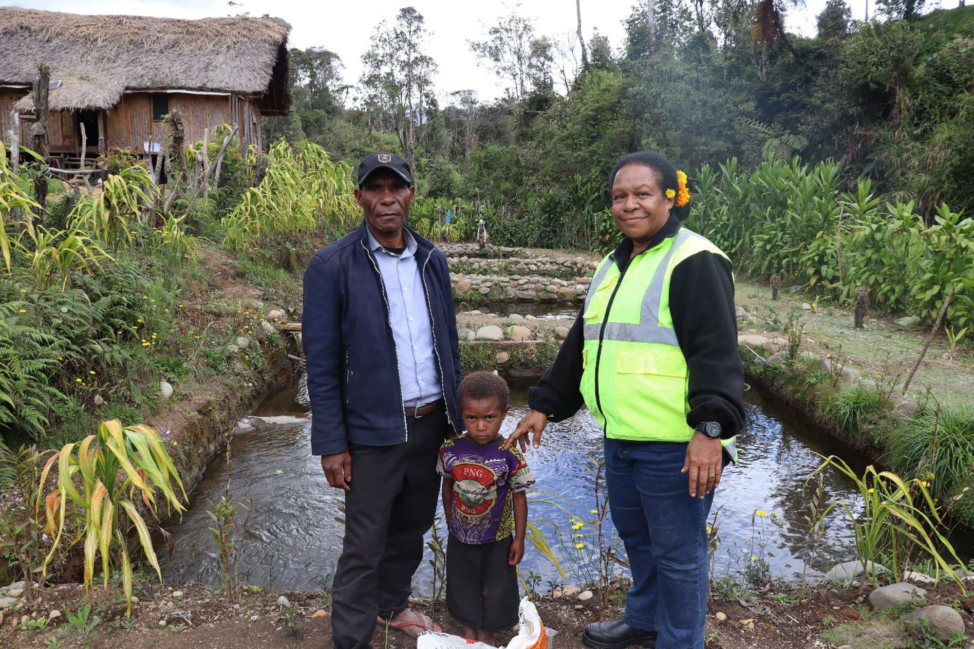Two adults and a child standing beside a small fish pond in a rural community.