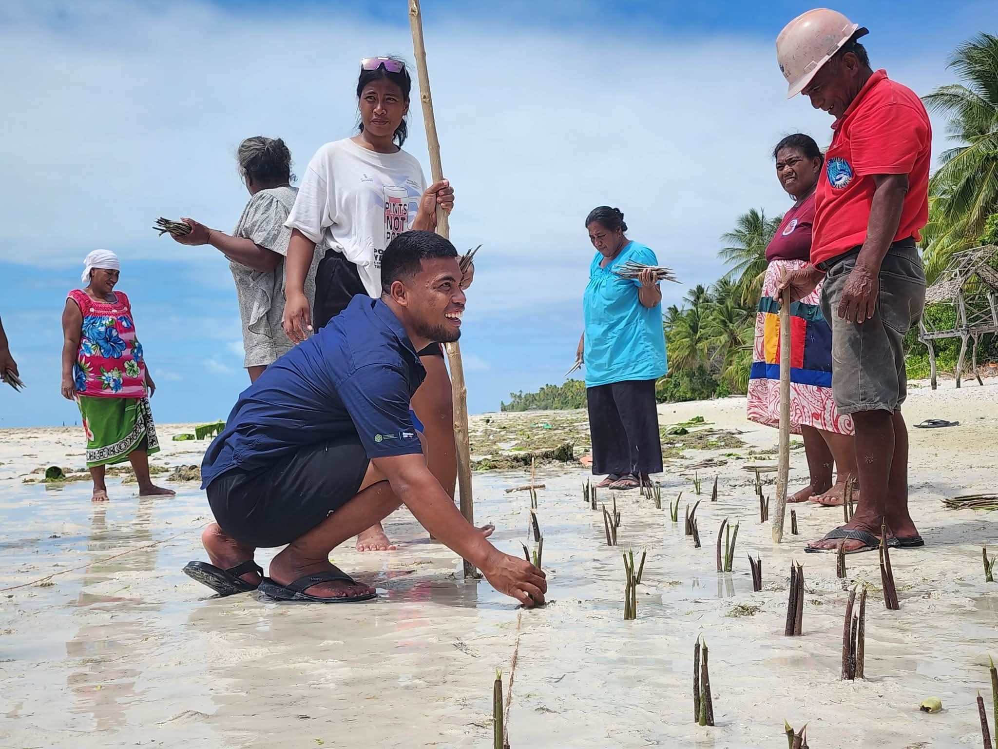 Community members plant mangrove seedlings along a sandy shoreline.