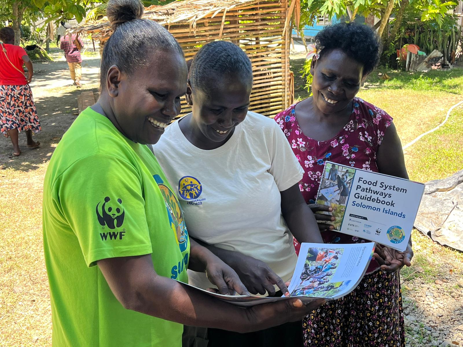 Three community members review a Food System Pathways Guidebook outdoors in the Solomon Islands.